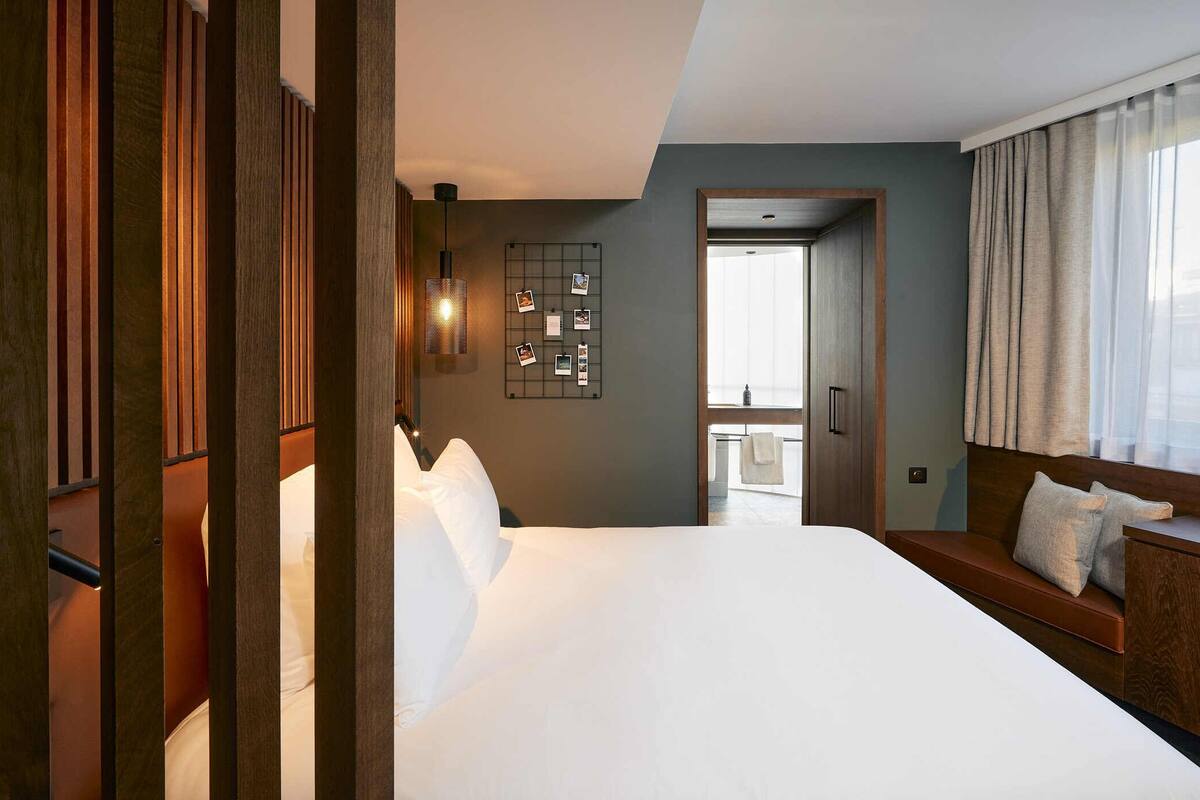 A hotel room with a large bed, a wooden headboard, a bedside table, a window with curtains, and a wall-mounted light fixture.