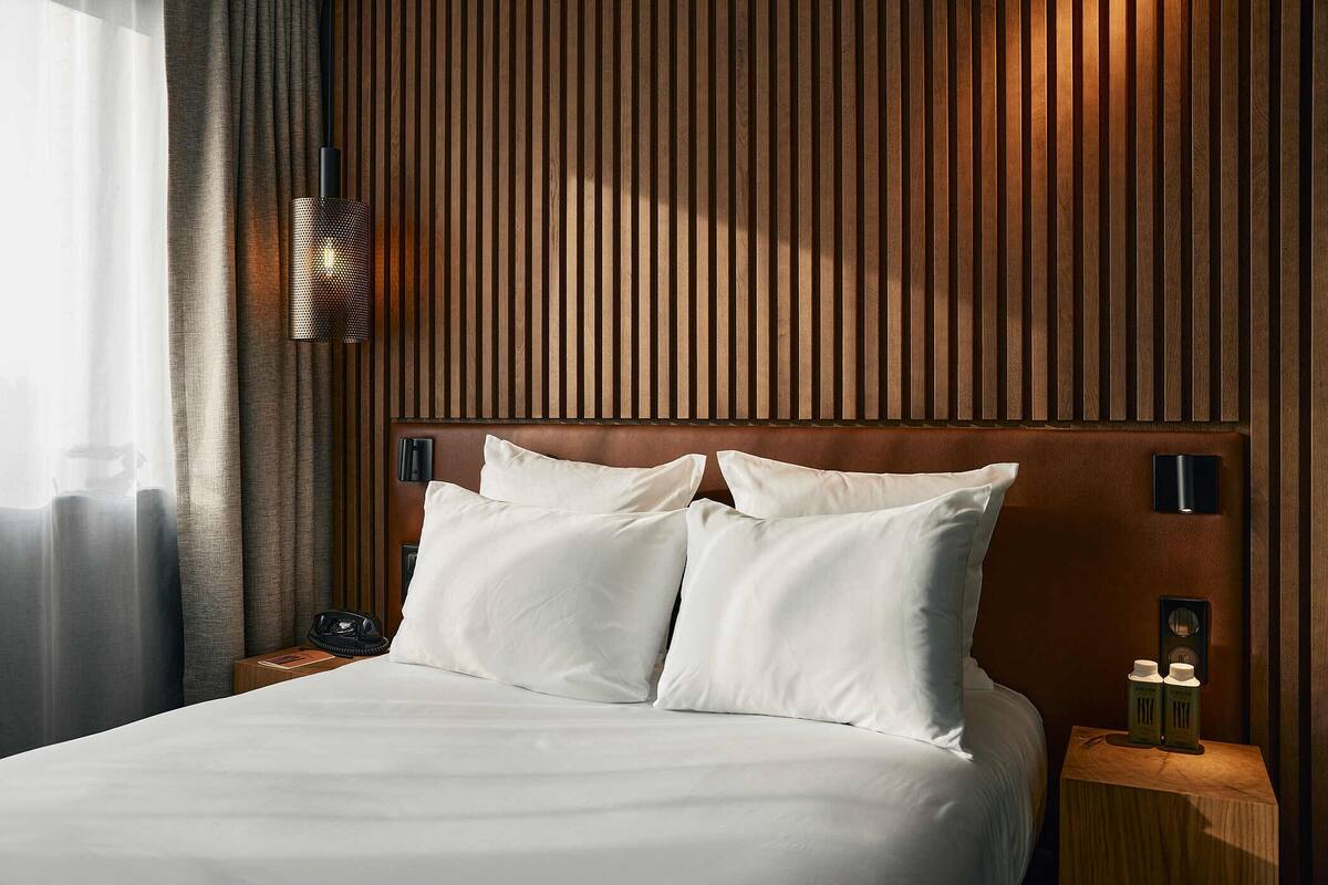 A hotel room with a wooden headboard, white bedding, a bedside lamp, and a nightstand with a phone and two bottles.