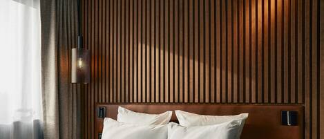 Habitación de hotel con cabecera de madera, ropa de cama blanca, lámpara de noche y mesita con un teléfono y dos botellas.