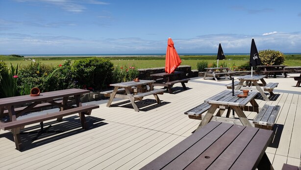 Eine Terrasse im Freien mit Holztischen und Sonnenschirmen, mit Blick auf eine Wiese und den Ozean.