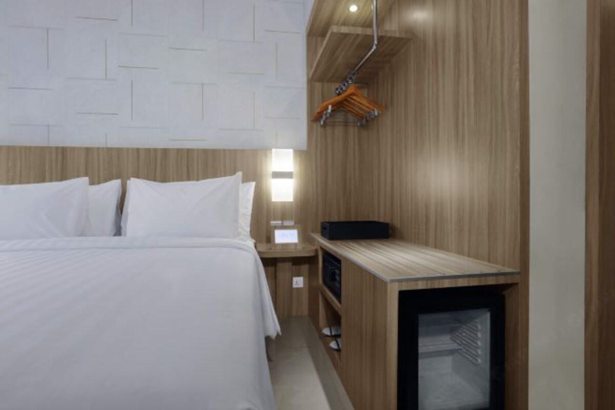 Habitación de hotel con cabecera de madera, cama con ropa blanca, mesita de noche con un objeto negro y armario de madera con espejo.