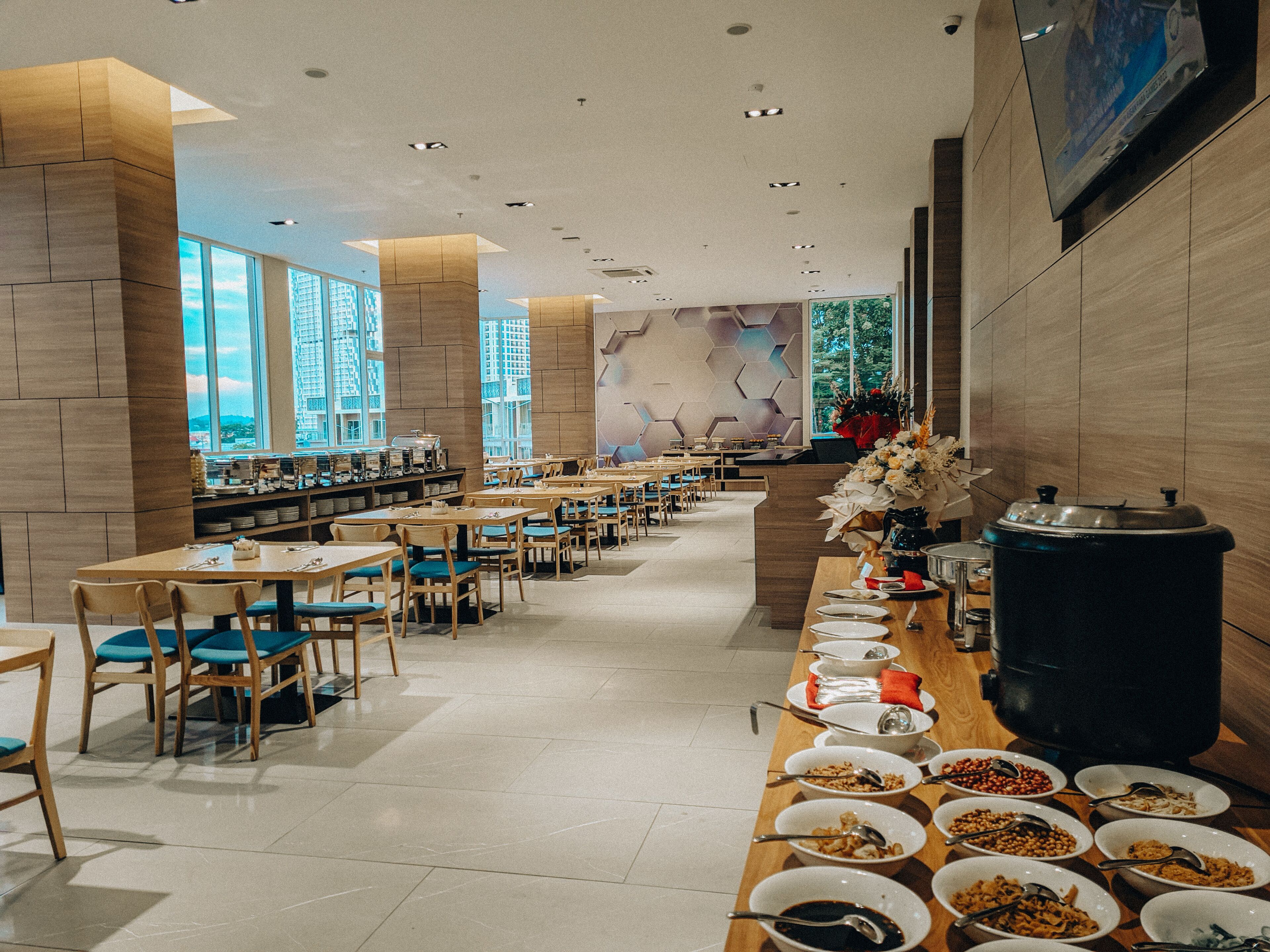 A spacious cafeteria with multiple tables and chairs, a buffet line with various food items, and large windows providing natural light.