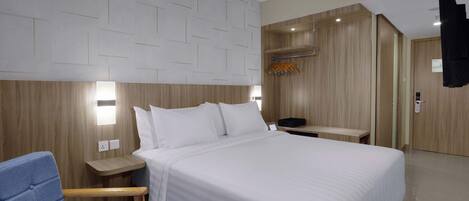 A hotel room with a large bed, a blue chair, a wooden nightstand, a wooden wardrobe, and an air conditioning unit.