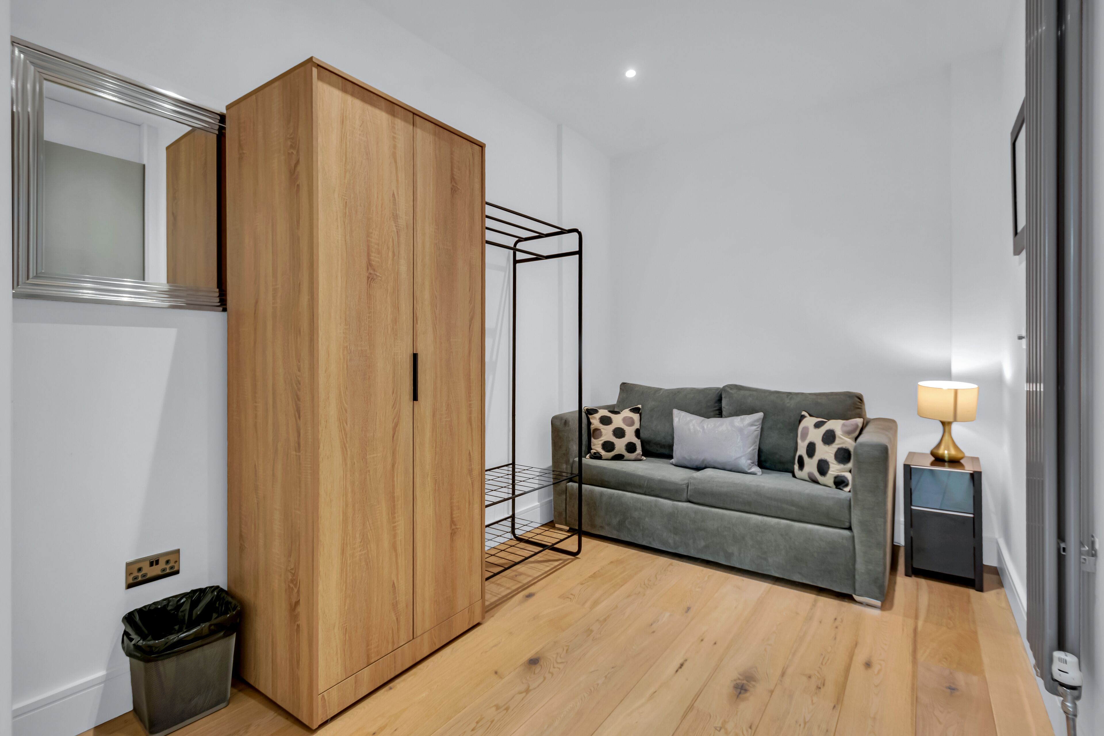 A room with a wooden wardrobe, a grey sofa with patterned cushions, a small table with a lamp, and a trash bin.