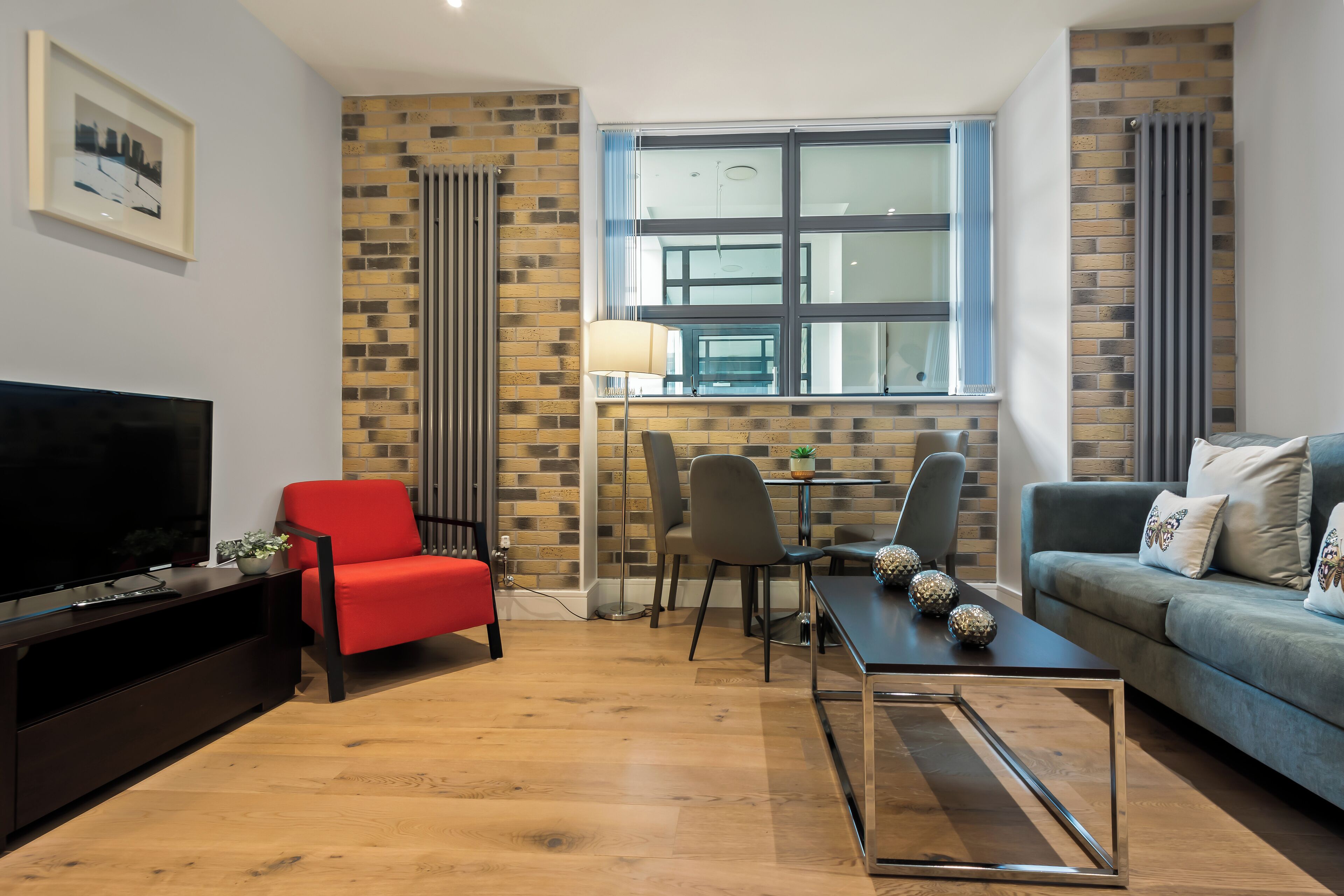 Uma sala de estar moderna com uma televisão de tela plana, poltronas vermelhas e cinzentas, um sofá azul e uma mesa de jantar com cadeiras.