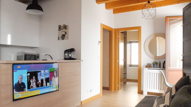 Ein modernes Wohnzimmer mit Flachbildfernseher, Holzboden, einer Couch und einer Küchenzeile mit Mikrowelle und Kaffeemaschine.
