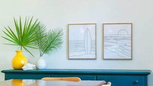Un coin repas avec une table et des chaises en bois, une commode bleu-vert et deux œuvres d’art encadrées représentant une planche de surf et un coucher de soleil sur l’océan.