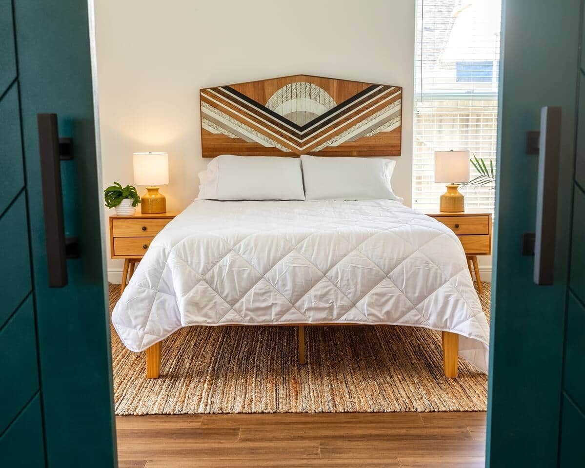 Een bed met een wit geborduurde sprei, een houten hoofdbord met een geometrisch ontwerp, twee nachtkastjes met lampen en een geweven vloerkleed op een houten vloer.