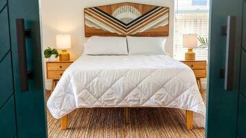 Een bed met een wit geborduurde sprei, een houten hoofdbord met een geometrisch ontwerp, twee nachtkastjes met lampen en een geweven vloerkleed op een houten vloer.