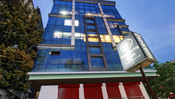 A modern hotel building with a glass facade, a sign indicating it is the HSB Hotelspring Brook, and red shutters on the ground floor. - Treebo Spring Brook (Guwahati)