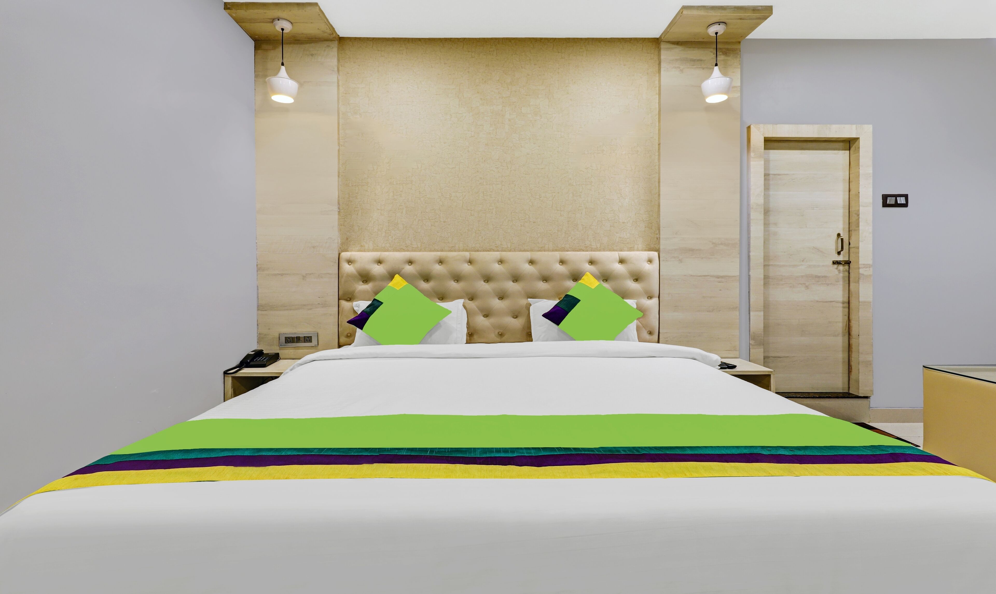 A hotel room with a tufted headboard, a bed with a colorful striped bedspread, two bedside lamps, and a door with a handle.