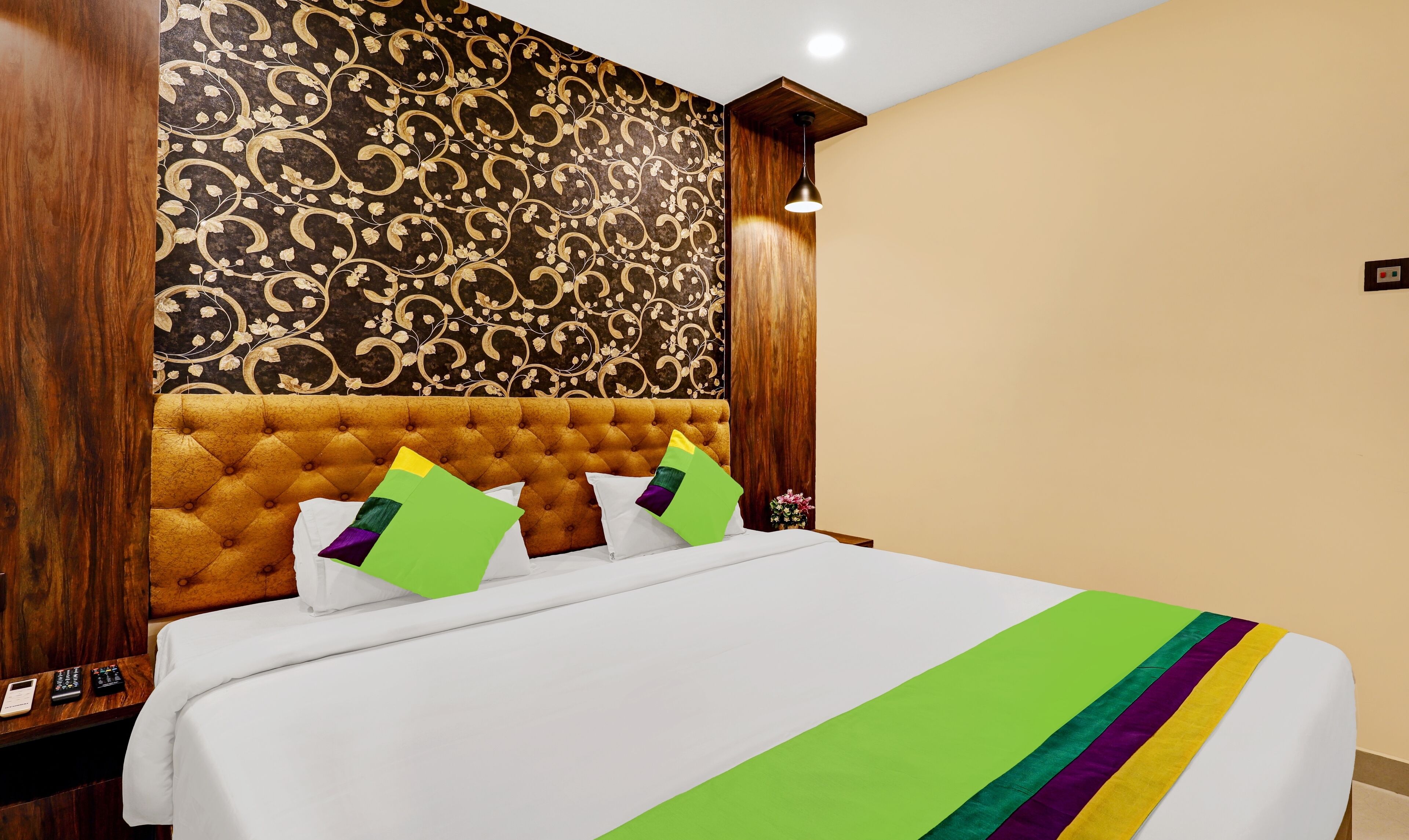 Een hotelkamer met een gestoffeerd hoofdeinde, een bed met wit beddengoed en een meerkleurige deken, en een wand met een bloemenprint.