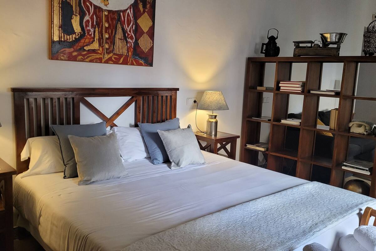 Un lit bien fait, avec une tête de lit en bois, deux lampes de chevet, une bibliothèque remplie de livres et d’objets décoratifs, et une peinture abstraite aux couleurs vives accrochée au mur.