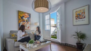 Una sala de estar con un sofá, una mesita de centro y dos personas sentadas conversando. Hay un ventanal con vistas a un edificio.