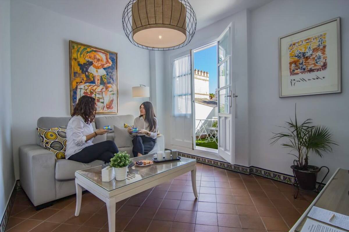 Un salon avec un canapé, une table basse et deux personnes assises, en train de discuter. Une grande fenêtre offre une vue sur un bâtiment à l’extérieur.