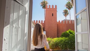 Vista desde un balcón con una puerta blanca, una persona de pie y un edificio rojo con torre y palmeras al fondo.