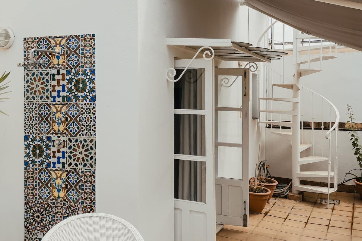 Une terrasse extérieure blanche, agrémentée d’un escalier en colimaçon blanc, de meubles en osier blanc et d’un mur recouvert de carreaux aux motifs géométriques.