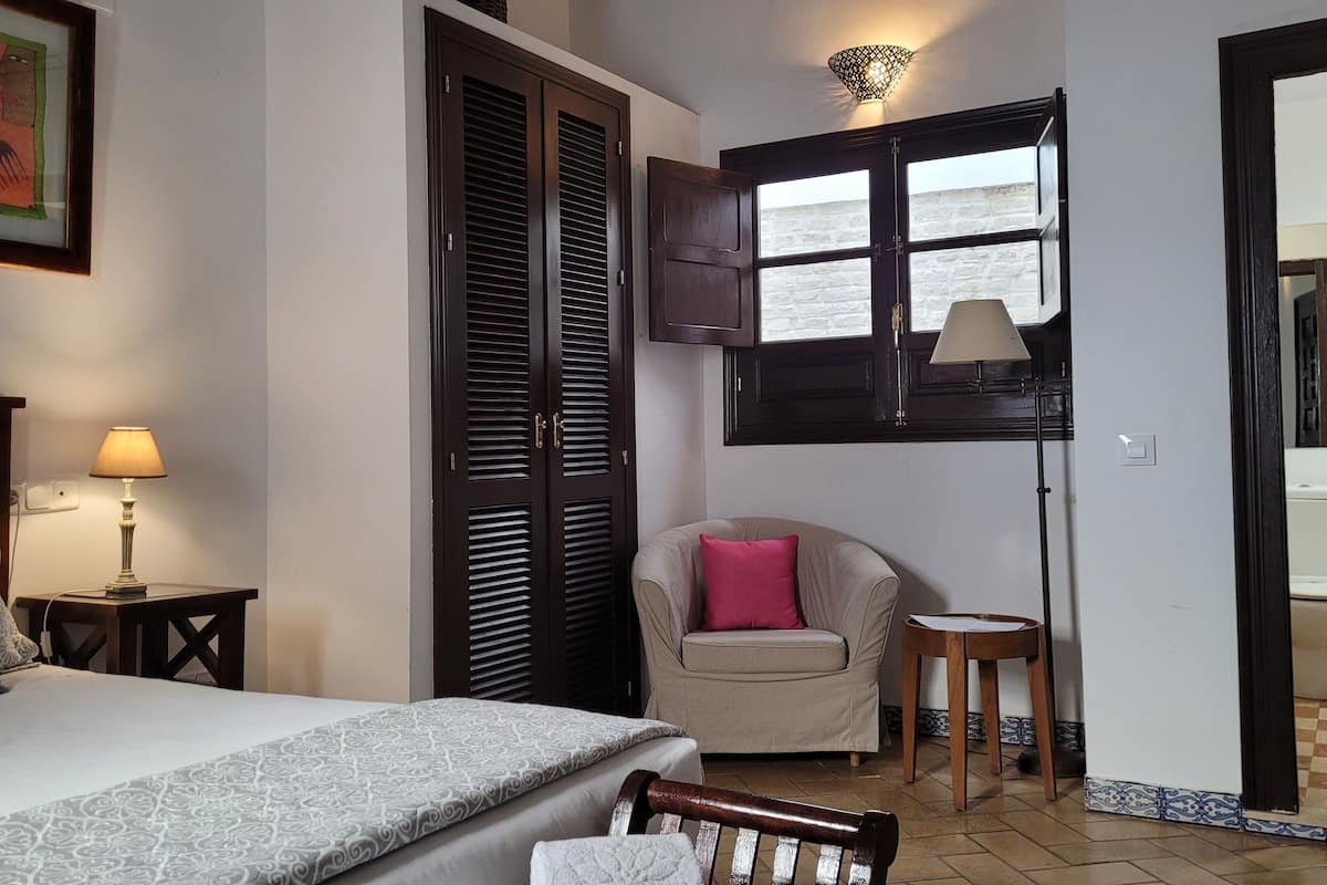 Une chambre à coucher comprenant un lit, une chaise, une table de chevet avec une lampe, une fenêtre avec des volets et une porte donnant sur une salle de bain.