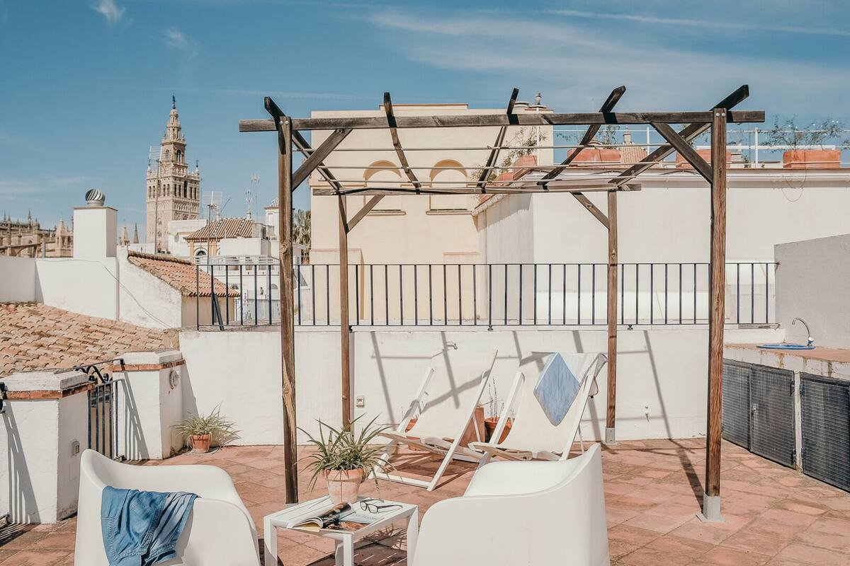 Une terrasse sur le toit agrémentée d’une pergola, de chaises longues blanches, d’une petite table avec une plante, et offrant une vue sur un bâtiment historique en arrière-plan.