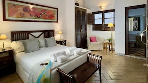 Un dormitorio con una cama grande, cabecera de madera, un banco, una mesita de noche con lámpara, un cuadro en la pared y una ventana con persianas.