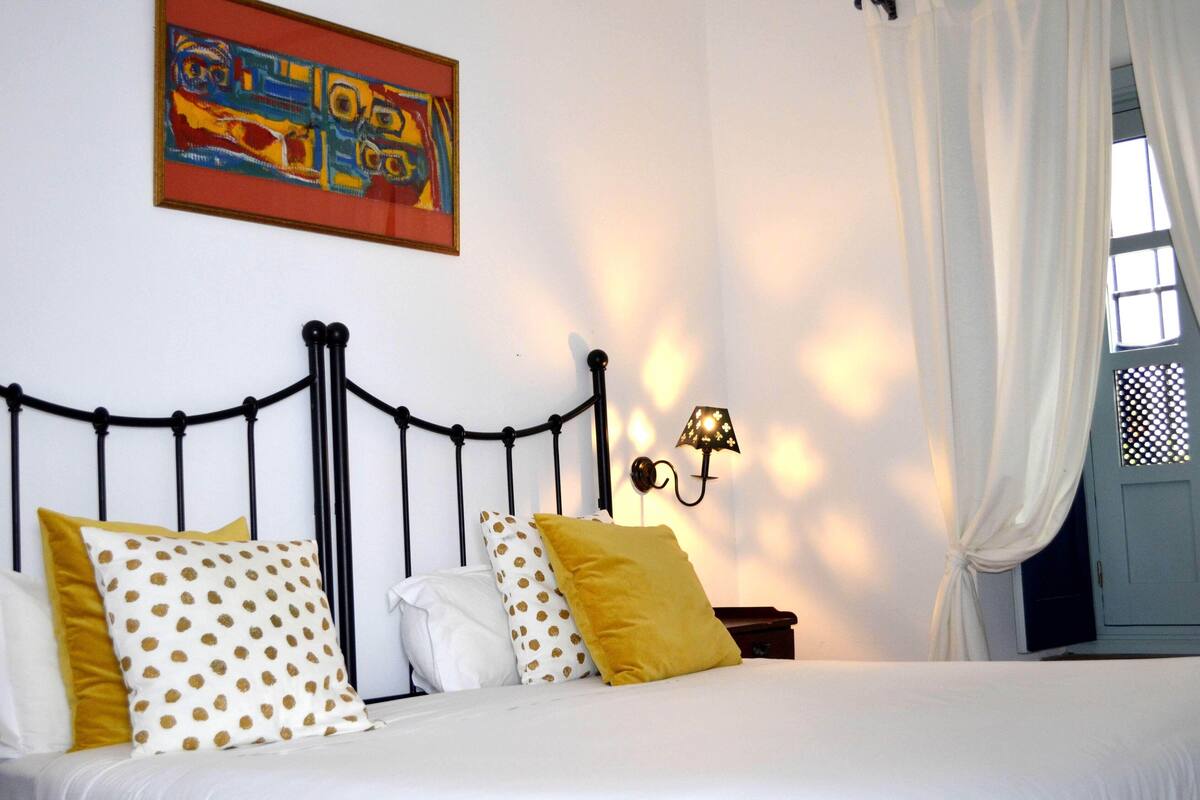 Un lit avec une literie blanche et plusieurs oreillers, une tête de lit en métal noir, une lampe fixée au mur et un tableau abstrait coloré au-dessus du lit.