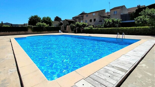 Une piscine rectangulaire bordée d’une terrasse en bois, entourée d’une haie basse et avec des immeubles d’habitation en arrière-plan.