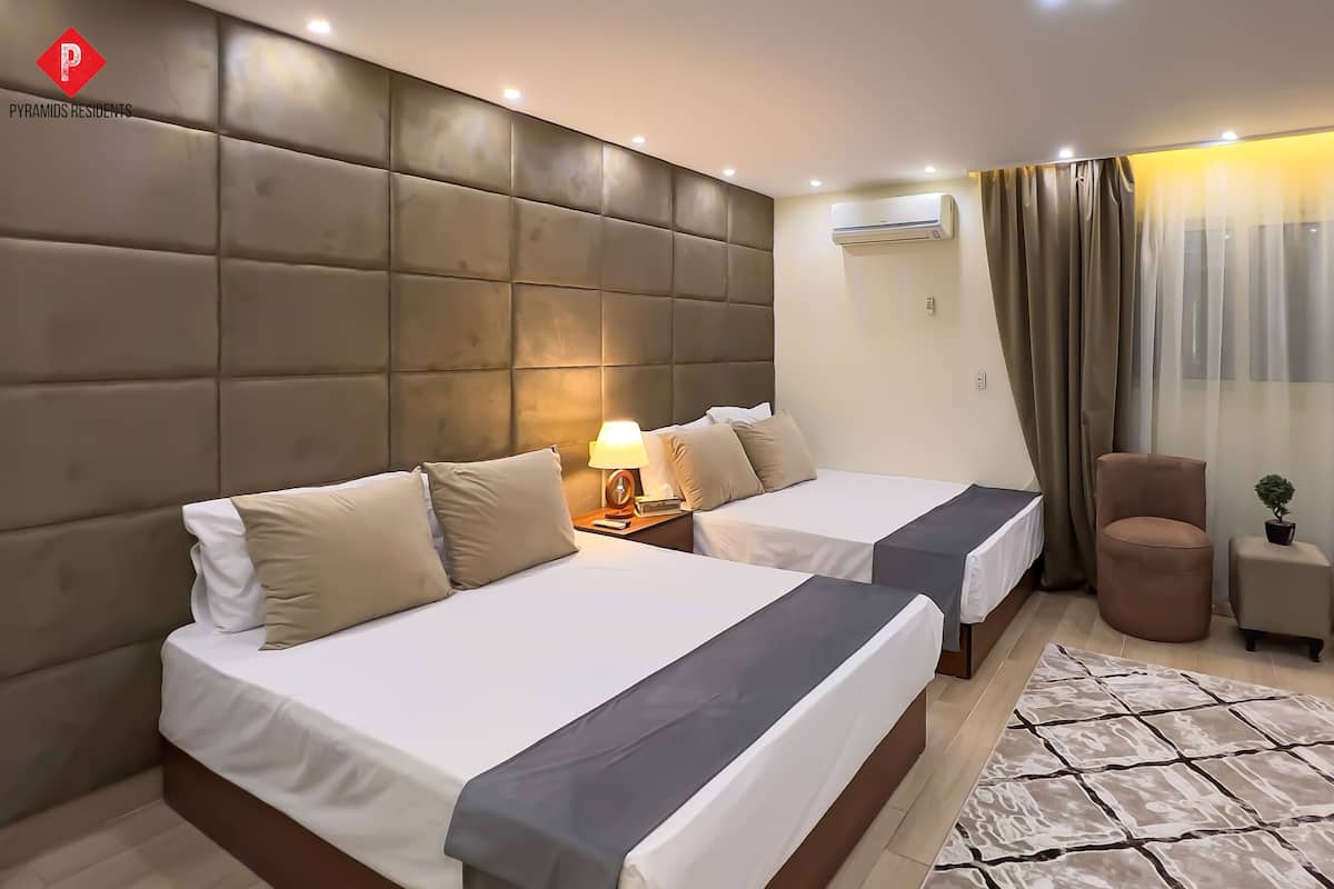 A hotel room with two beds, a padded headboard, an air conditioning unit, a lamp, a small table with a plant, and a patterned rug.