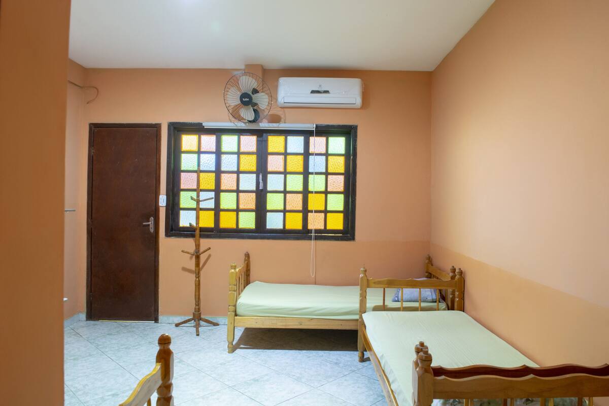 A bedroom with two wooden beds, a window with colorful glass panes, a standing fan, and an air conditioning unit on the wall.
