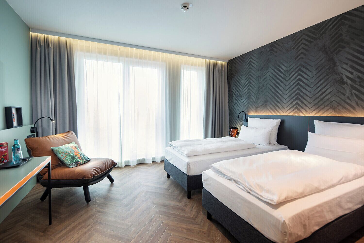 A hotel room with two beds, a desk, a chair, and a large window with curtains.