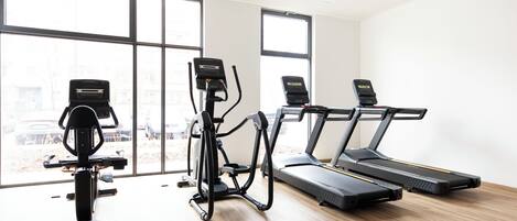 Un gimnasio con equipo cardiovascular, incluyendo cintas de correr y una máquina elíptica, con grandes ventanales y vistas a coches estacionados afuera.