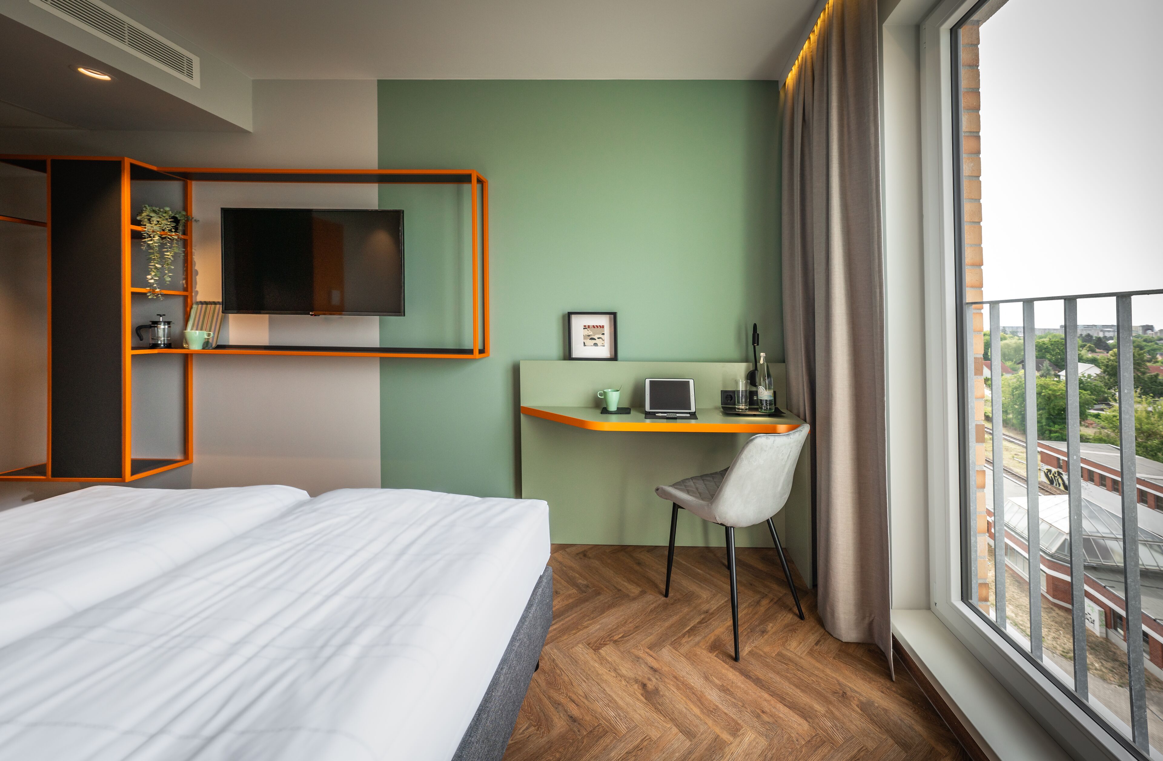 Ein Hotelzimmer mit einem Bett, einem Schreibtisch mit Laptop, einem Fernseher und einem Fenster mit Blick auf Gebäude.
