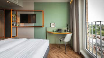 Ein Hotelzimmer mit einem Bett, einem Schreibtisch mit Laptop, einem Fernseher und einem Fenster mit Blick auf Gebäude.