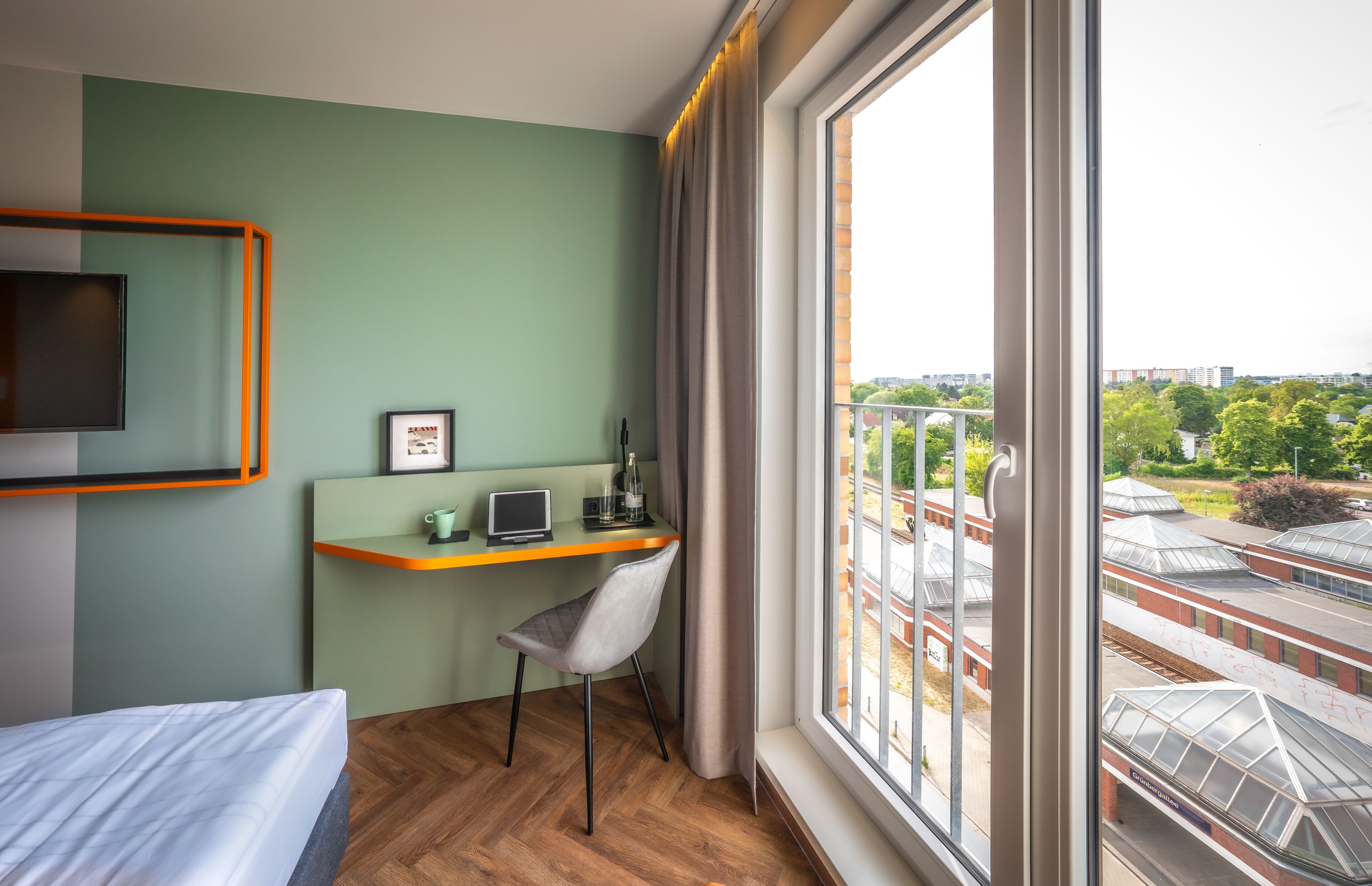 Habitación de hotel con cama, escritorio con silla, televisión y ventana grande con vista a la ciudad.