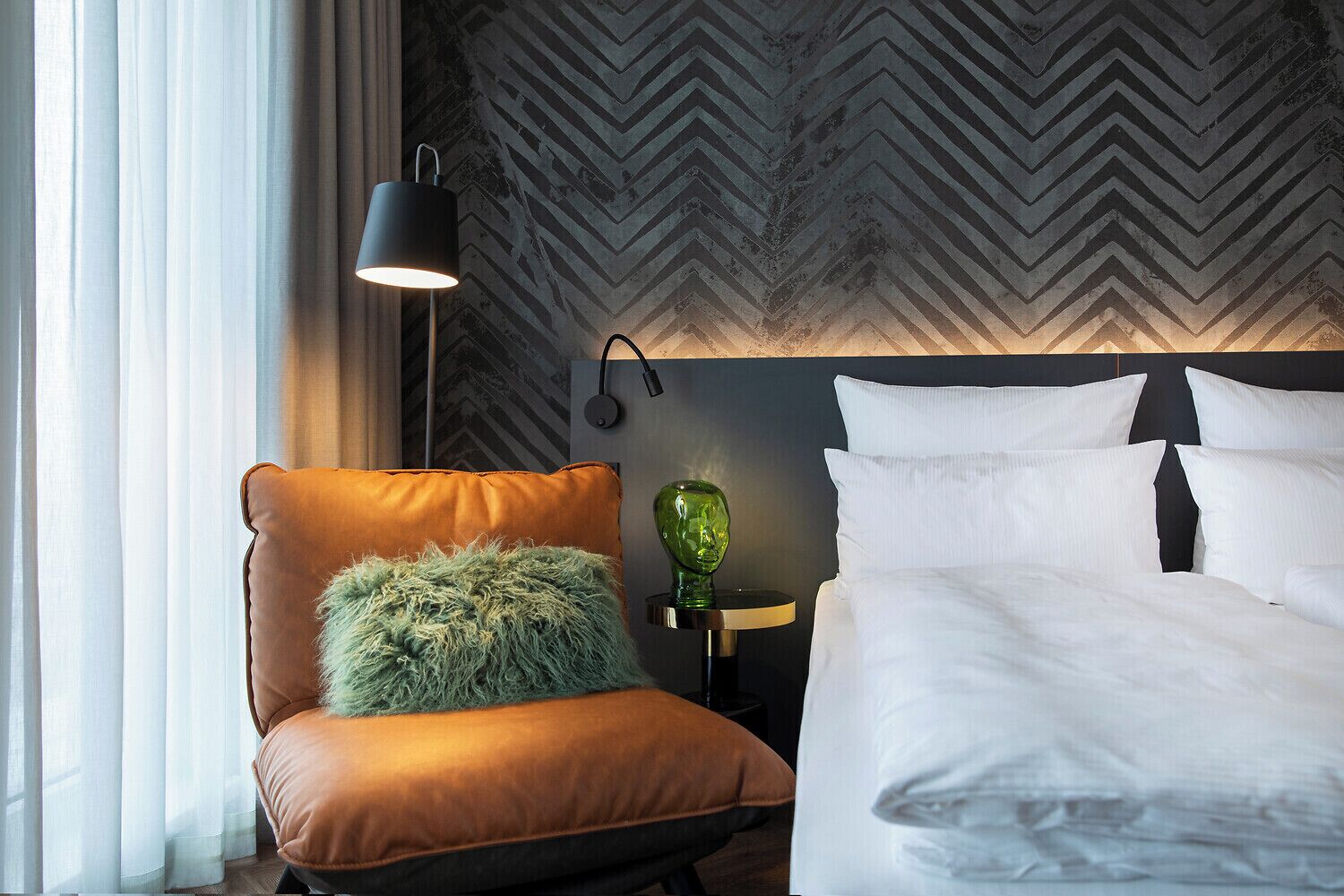 Ein Hotelzimmer mit Bett, Sessel mit flauschigem Kopfkissen, Lampe und Wand mit Zickzackmuster.