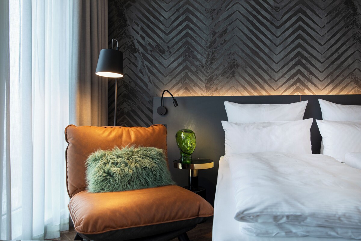 Une chambre d’hôtel moderne avec un grand lit, un fauteuil en cuir, un vase vert et une lampe fixée au mur.