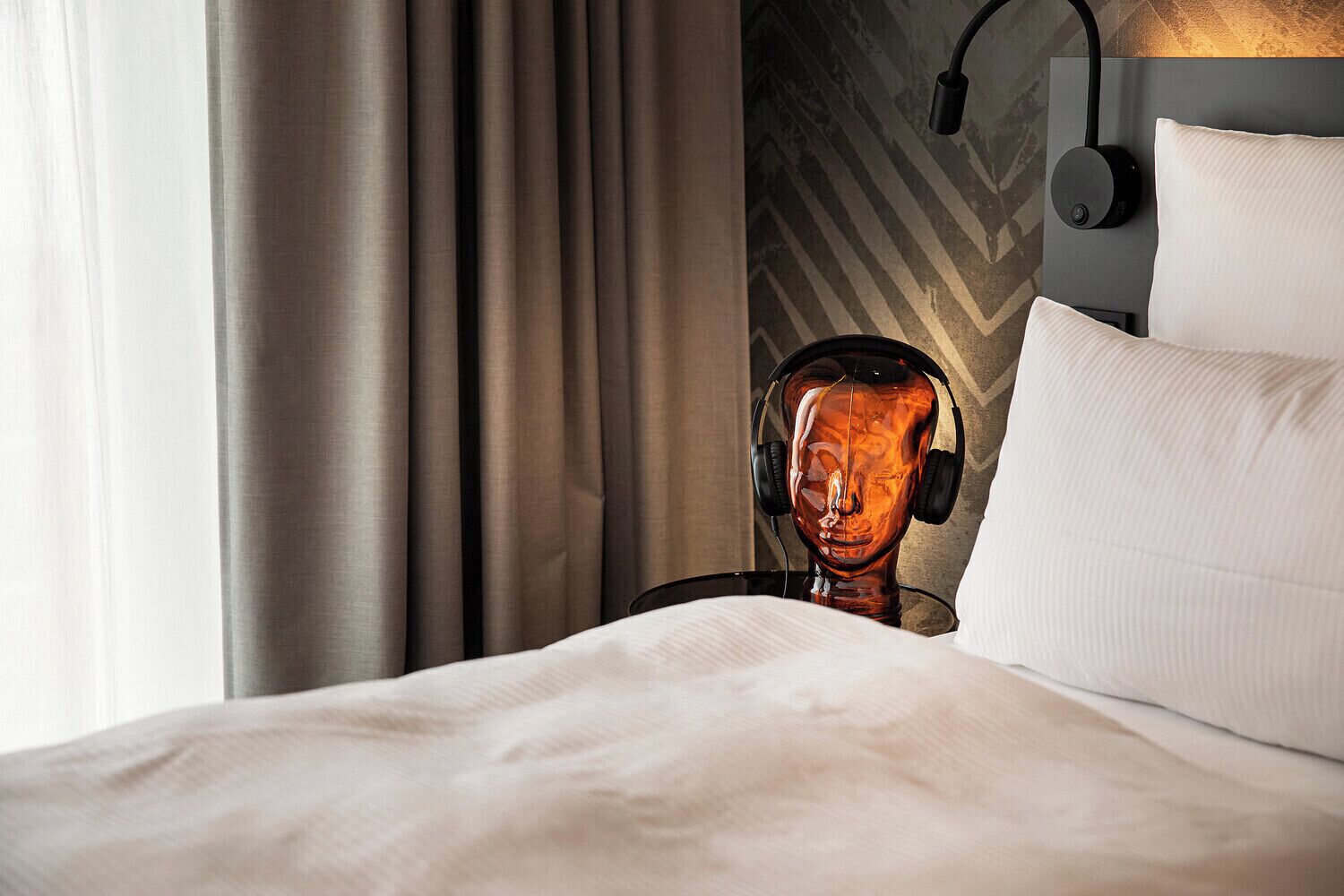 A hotel room with a bed, a headboard with a geometric pattern, a wall-mounted lamp, and a window with curtains.
