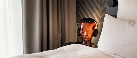 A hotel room with a bed, a headboard with a geometric pattern, a wall-mounted lamp, and a window with curtains.