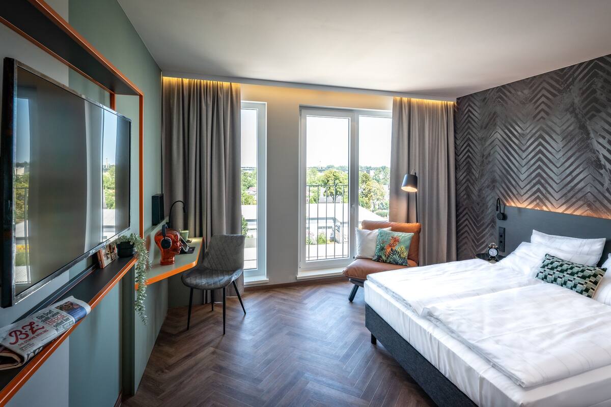 Een hotelkamer met een groot bed, een bureau met een computer, een stoel en een televisie. Er is een balkon met gordijnen en uitzicht op de omgeving.