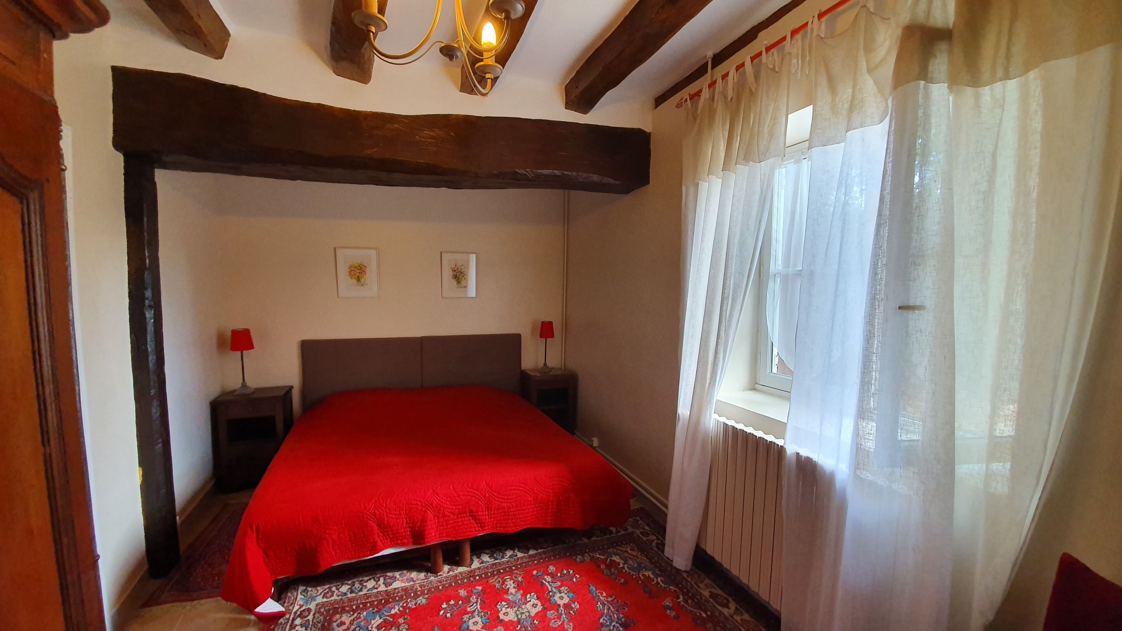 Ein Schlafzimmer mit einem großen Bett, Holzbalken, zwei Nachttischen mit Lampen, einem Fenster mit durchscheinenden Vorhängen und einem gemusterten Teppich.