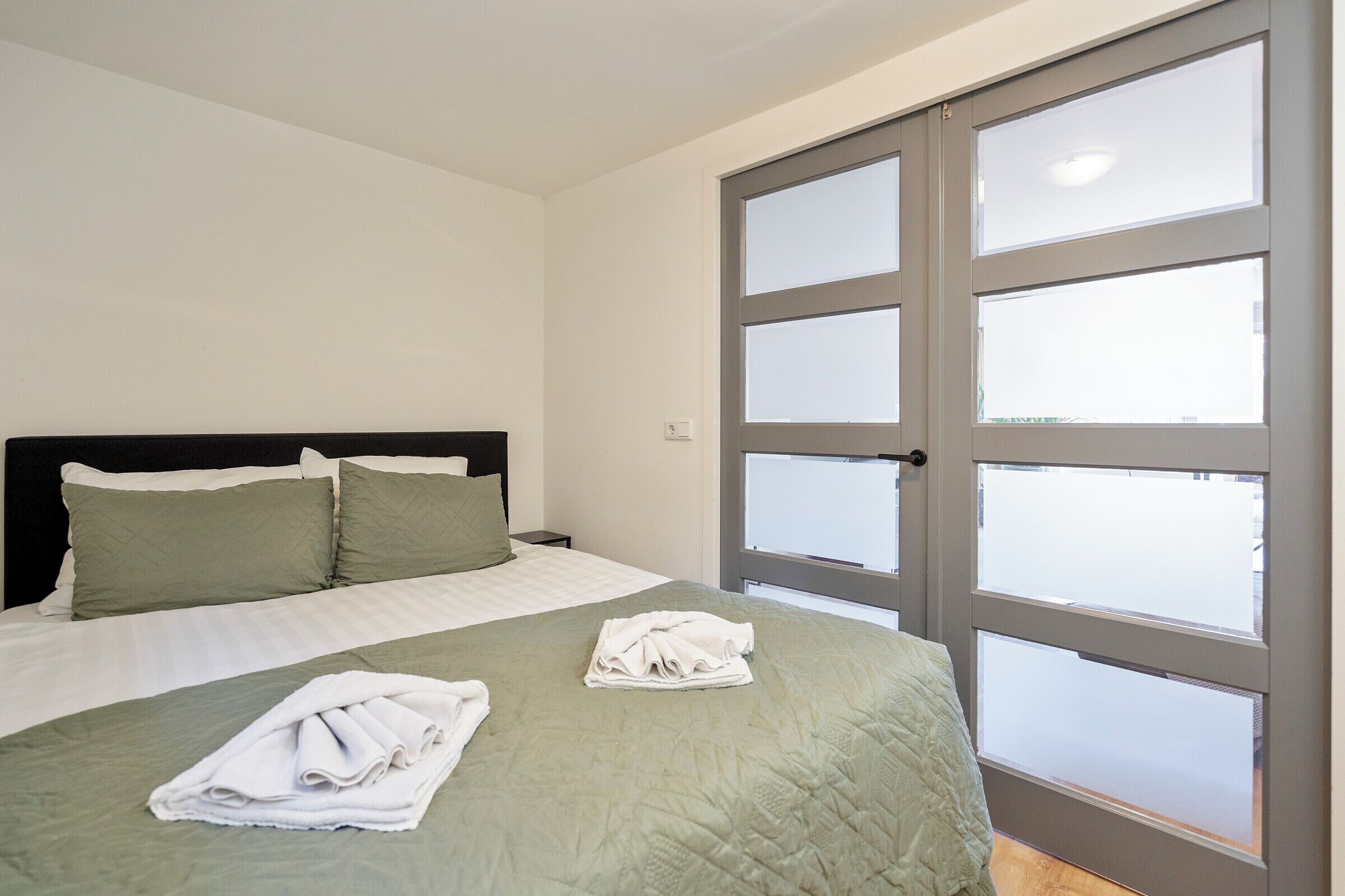Habitación de hotel con cama, dos almohadas, una toalla doblada y una puerta con paneles de vidrio esmerilado.
