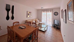 A dining area with a wooden table and chairs, a sofa, and a TV in the background. - Elegant terraced house in a fantastic south position in Els Poblets (Denia)