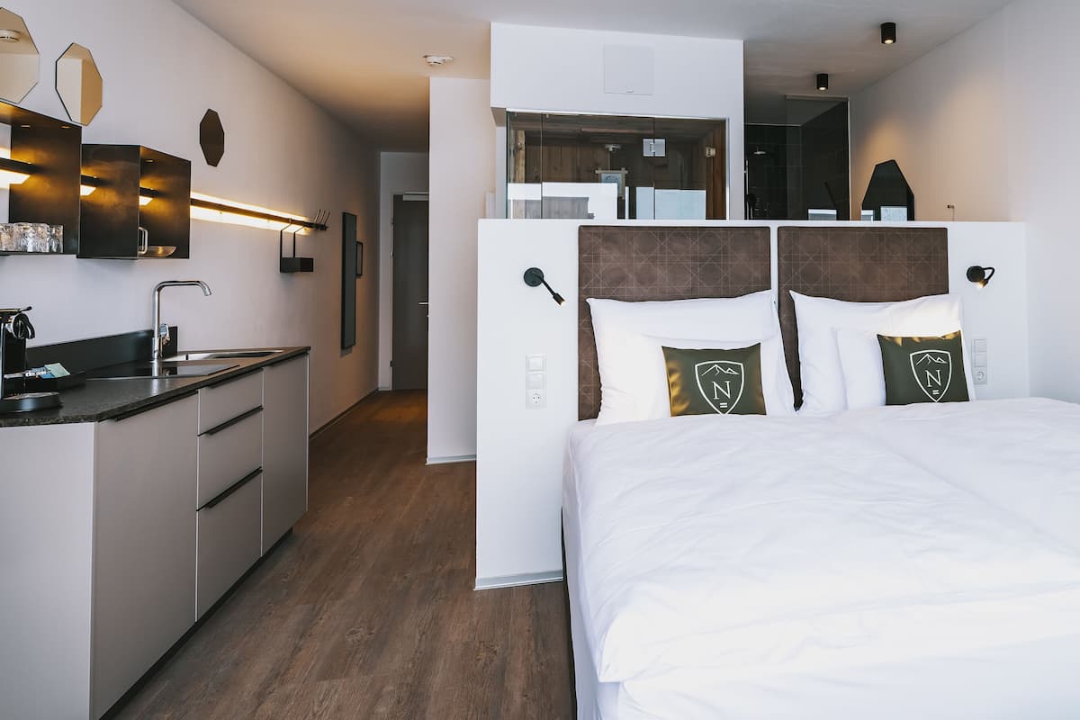 A modern hotel room with a bed, a kitchenette, and a bathroom visible through a glass partition.
