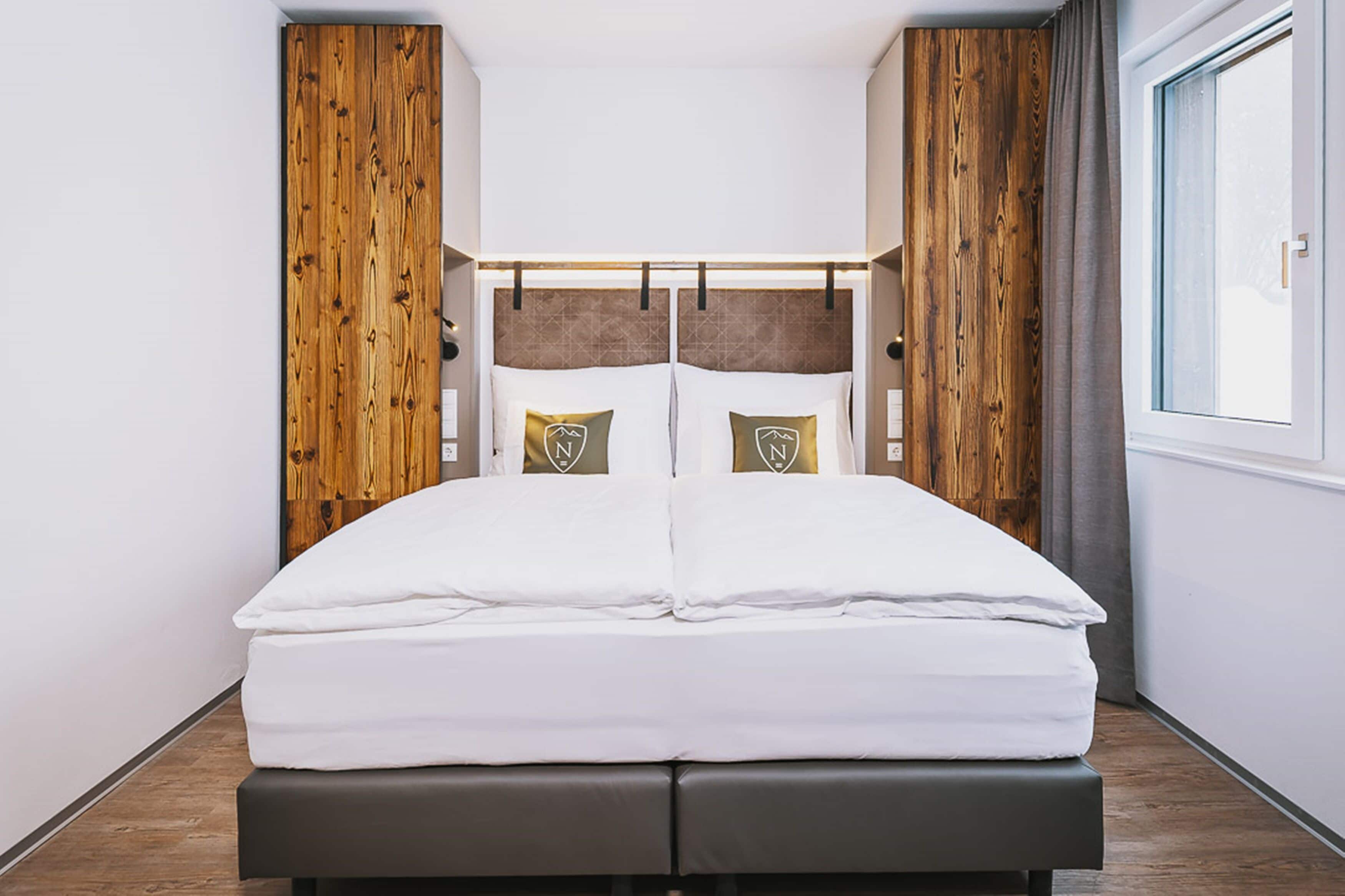 A double bed with a wooden headboard, two pillows with a logo, and a window on the right.