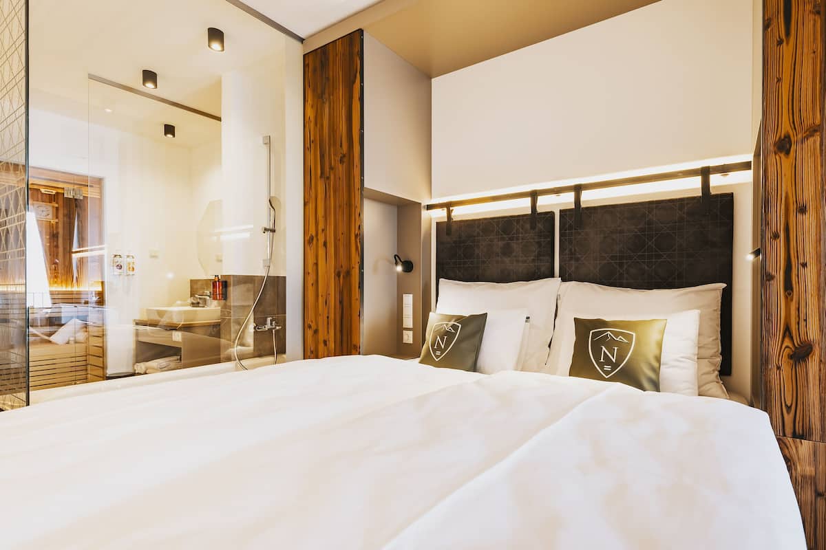 A hotel room with a large bed, wooden paneling, a bathroom with a glass shower enclosure, and a visible minibar.