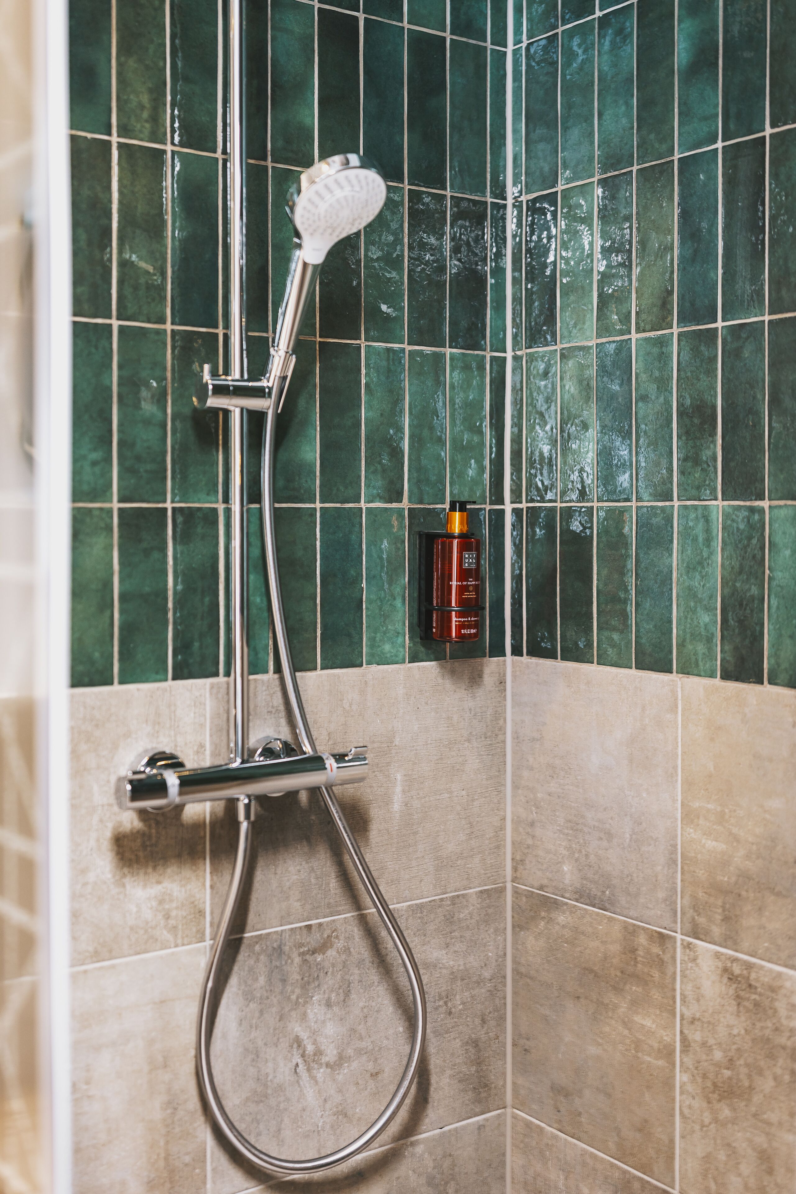 A shower with a handheld showerhead and a wall-mounted control panel, set against a tiled wall with a green and beige color scheme.