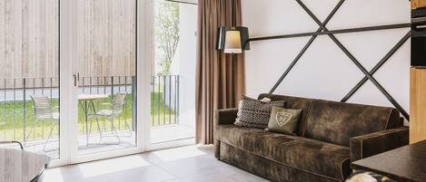 Ein modernes Wohnzimmer mit einer Couch, einem Tisch und einer Lampe. Eine Glastür öffnet sich zu einem Außenbereich mit Stühlen.