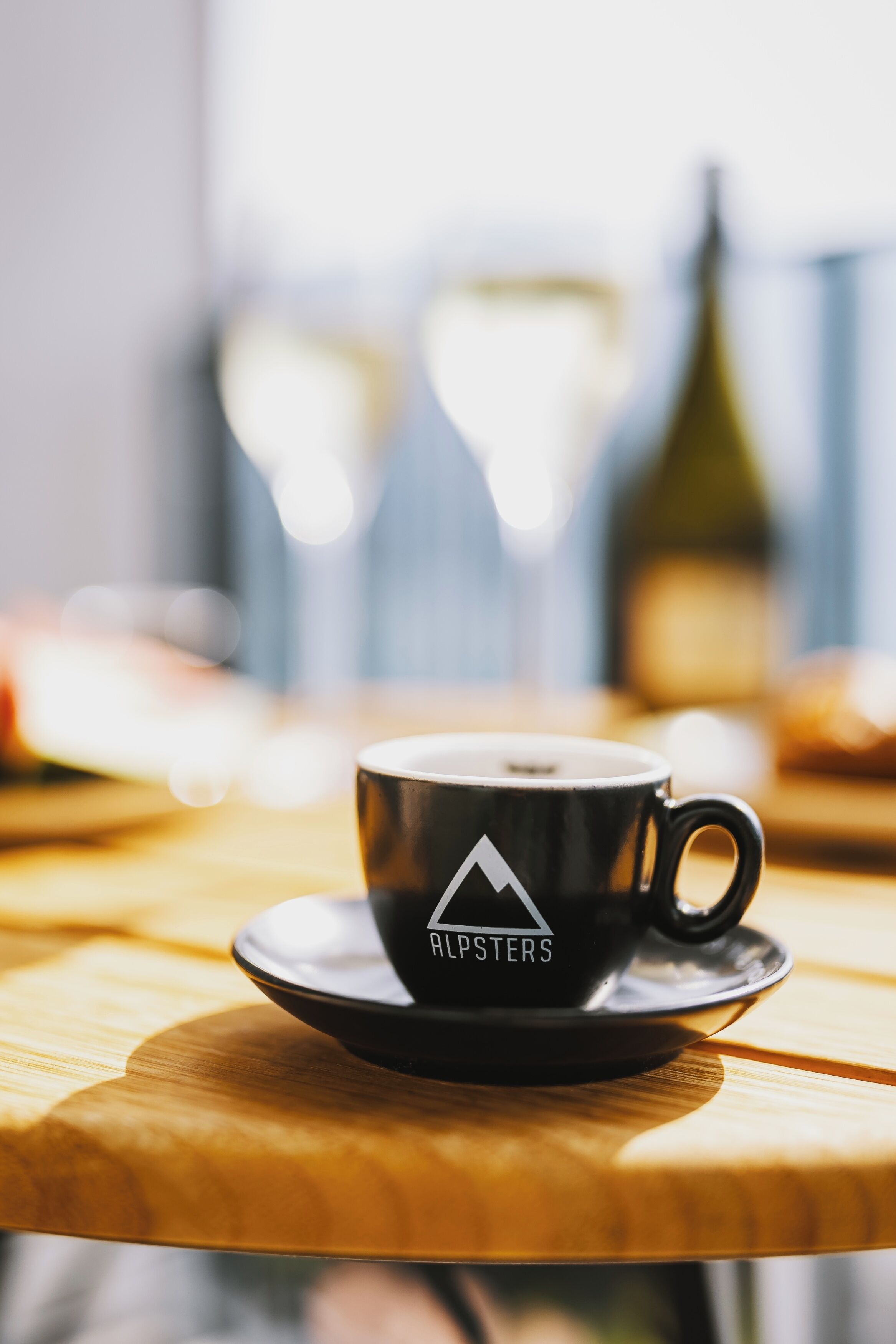 A black coffee cup with a mountain logo on a saucer, placed on a wooden table with a blurred background.