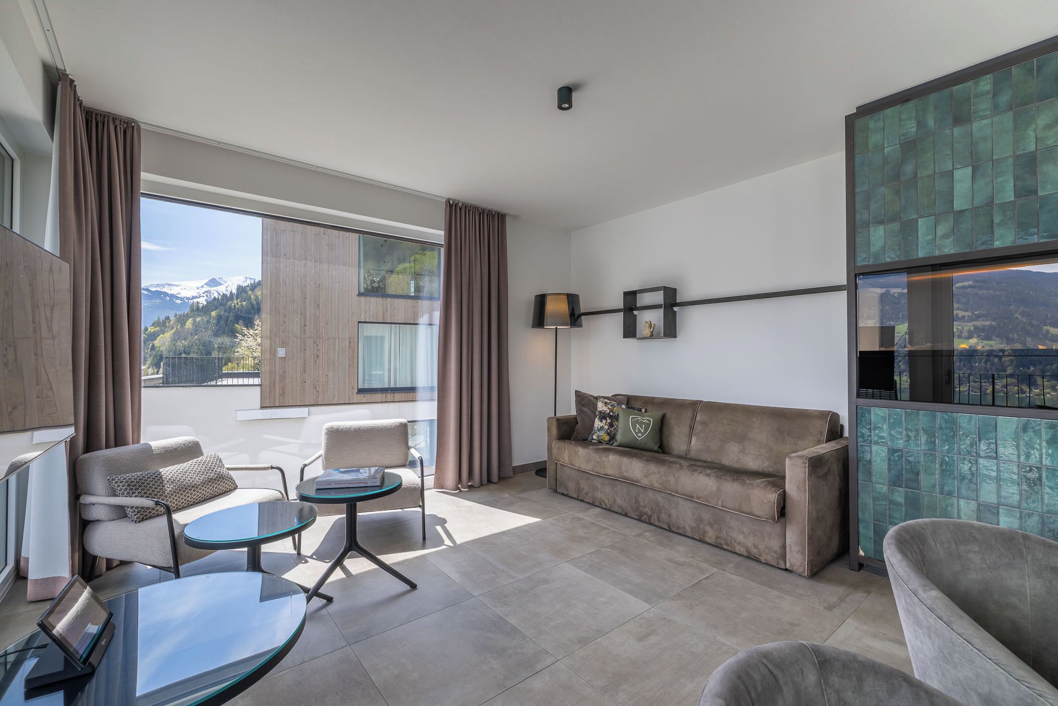 Un salon moderne avec un canapé, des fauteuils, une table en verre et une vue sur les montagnes, visible à travers de grandes fenêtres.