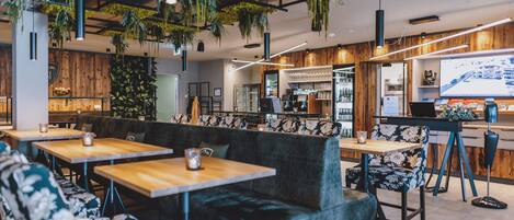 A modern cafe with wooden tables, cushioned seating, hanging plants, and a bar area.