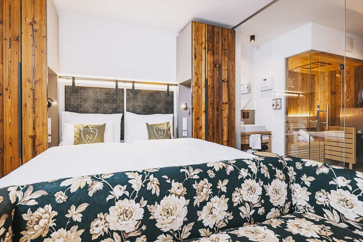 A modern hotel room with a large bed, wooden accents, a floral sofa, and a bathroom with a glass enclosure.